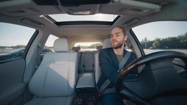 Caucasian handsome man with brown hair in bun driving car, holding steering wheel with both hands, turning head to left. Male wearing dark blazer and green shirt. Sunroof open, empty passenger seat.
