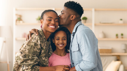 A family stands together in their living room. One adult gives a kiss on the forehead of the other while they all smile. The setting is warm and inviting. © Prostock-studio