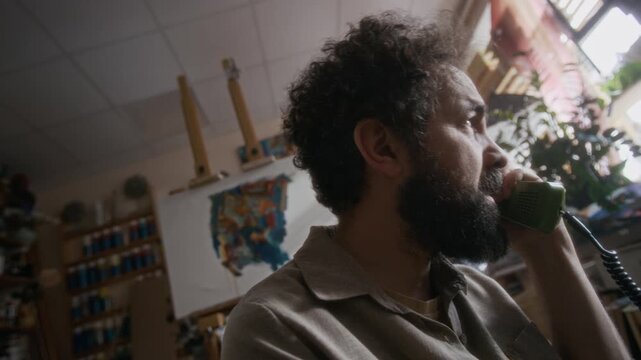 Dutch angle close up shot of desperate male painter sitting in his workshop, having difficult conversation on landline phone, then covering face with hands while feeling sad and hopeless