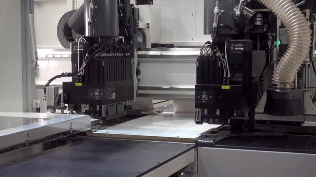Automated machine heads move over a flat white panel in an industrial factory, cutting or processing sheet material with precision in 4K.