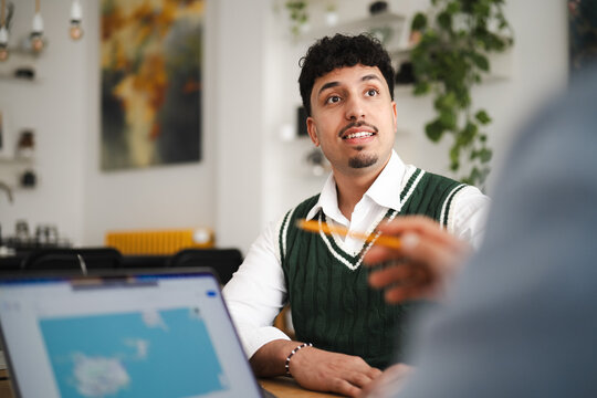 A young man in a green sweater vest and white shirt engages in a discussion with a colleague, holding a pen and looking towards a laptop screen.