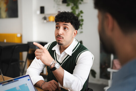 A young man in a white shirt and green sweater vest gestures while speaking to another person across a table with a laptop.