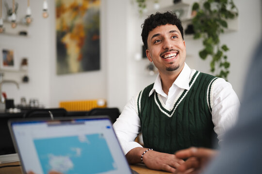 A young man smiles while looking up during a meeting. He is wearing a white shirt and a green sweater vest.