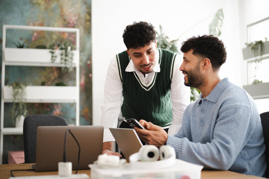 Two men collaborate on a project, reviewing information on a phone and laptop in a modern office setting with plants.