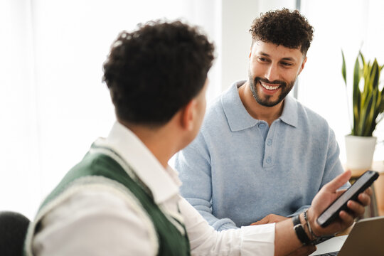 Two men are in a bright room, one holding a smartphone and smiling while talking to the other.