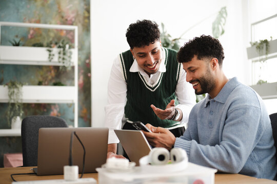 Two young men collaborate on a project, one showing the other something on a mobile device while laptops are on the table.