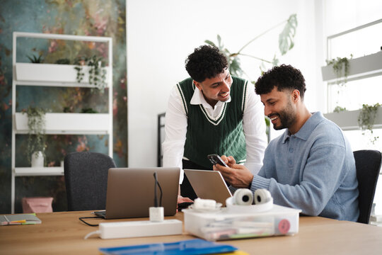 Two men collaborate at a desk, one showing the other something on his phone while a laptop is open.