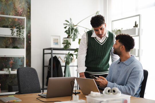 Two young men collaborate at a desk with laptops and a smartphone, discussing ideas in a modern office setting with plants.
