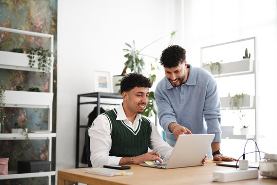 Two young men collaborate on a laptop at a desk, discussing ideas in a bright, modern office space with plants.