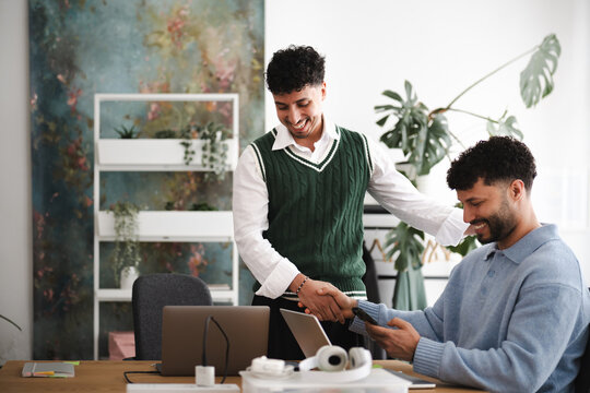 Two men shake hands in an office setting, one holding a phone and the other smiling, signifying a successful business agreement or partnership.