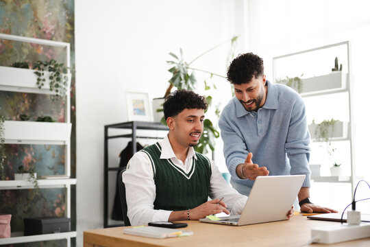 Two young men collaborate on a project, one seated at a laptop and the other standing and gesturing, in a bright, modern office space.
