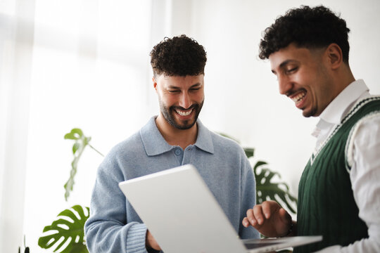 Two young men share a laugh while looking at a laptop screen, suggesting a moment of shared success or humor in a modern office setting.