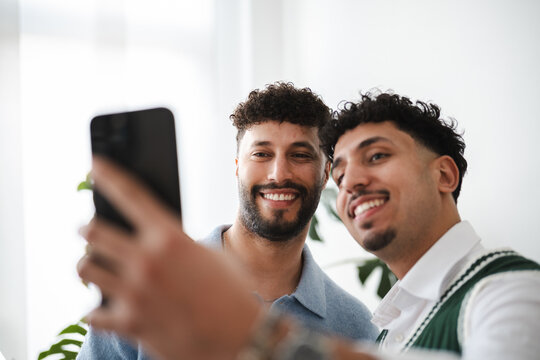 Two young men smile as they take a selfie with a smartphone, capturing a happy moment together indoors.