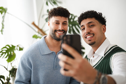 Two smiling young men take a selfie with a smartphone, surrounded by lush green plants in a bright, modern setting.