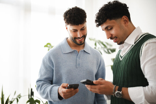 Two young men are looking at their phones and smiling. They are standing indoors with plants in the background.