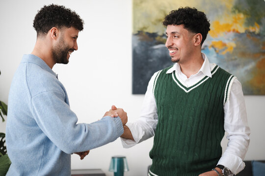 Two men shake hands, smiling warmly at each other in a modern office setting. This gesture signifies agreement, partnership, or a friendly greeting.