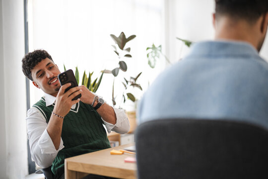 A man smiles while looking at his phone in an office setting. Another person is seated in the foreground, with their back to the camera.