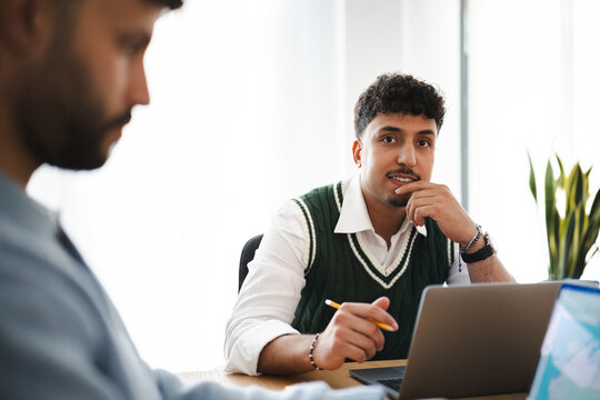Two colleagues collaborate on a project, one man looks thoughtfully at the camera while the other is out of focus, working on a laptop.