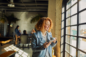 Fototapeta premium Smiling young woman with coffee and smartphone by office window in modern coworking space