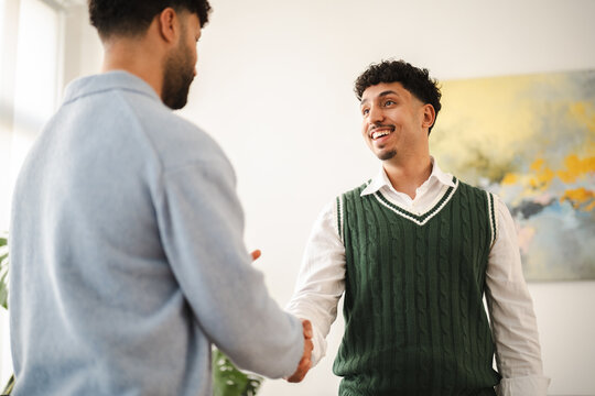Two men shake hands, one in a light blue sweater and the other in a white shirt and green sweater vest, smiling warmly in a bright, modern setting.