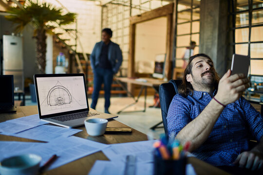 Architect checking smartphone beside laptop in loft office