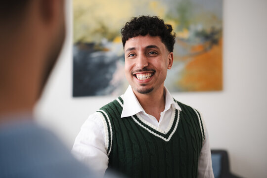 A man in a white shirt and green sweater vest smiles while talking to someone off-camera in an office setting.