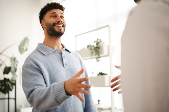 A man with a beard smiles and extends his hand to greet another person in a bright, modern office setting with plants.