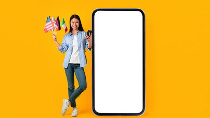 A young woman stands beside a large, empty screen while holding flags from different countries in one hand and a smartphone in the other. She is smiling and appears engaged in her activity. © Prostock-studio