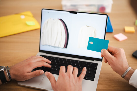 A person uses a laptop to shop online, holding a debit card to complete the purchase of clothing.