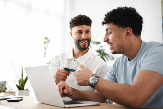 Two men are smiling while one hands a credit card to the other, who is using a laptop for an online transaction.