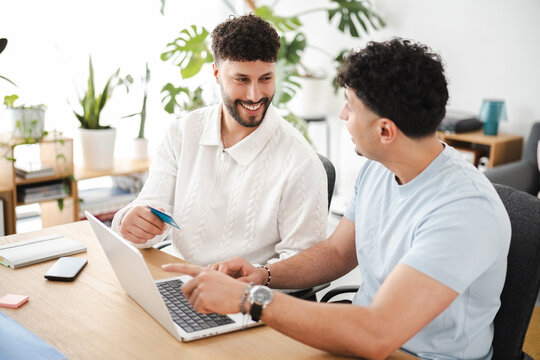 Two men are sitting at a desk, one holding a credit card and looking at a laptop while the other points at the screen.