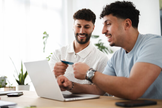Two men are smiling while one holds a credit card and the other looks at a laptop, suggesting online shopping or a financial transaction.