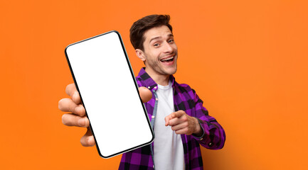 A young man stands in front of an orange wall. He holds up a smartphone in one hand and points towards the viewer with a smile. His expression shows excitement and friendliness. © Prostock-studio