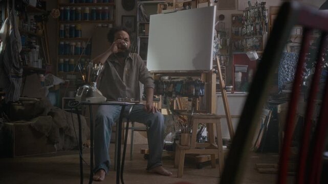 Handheld shot of stressed artist sitting drinking whiskey from glass, then standing up, picking palette and brush and going to paint on canvas in cluttered workshop