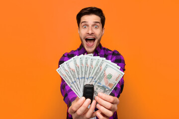 A man stands against an orange background and shows several hundred dollar bills and a car key. He looks very happy and is smiling widely while holding the money. © Prostock-studio