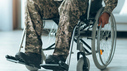 A veteran sits in a wheelchair while wearing camouflage pants. The setting is indoors, with light streaming in. The individual appears focused as they maneuver the wheelchair. © Prostock-studio