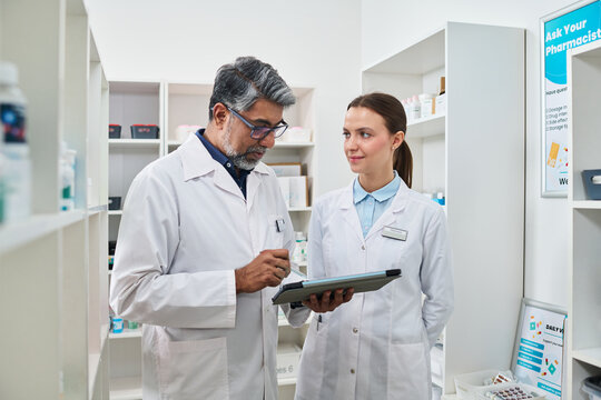 Middle aged Indian man and young woman reviewing tablet in pharmacy, coordinating medication workflow and patient service. For healthcare staffing or retail pharmacy marketing