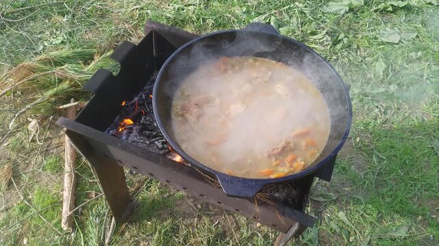 There is a metal brazier on the grass, in which firewood is burning. There is a metal cauldron on top of it, in which meat and vegetables are stewed. The cooking dish boils, gurgles and steam rises