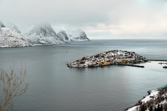 A view of a small island Husoy in Norway's waters surrounded by snow-covered hills. The sky is gray, and boats can be seen near the island. It is winter and very cold.