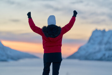 A woman stands with arms raised at sunset in Norway. The colorful sky reflects on the water, while snow-capped mountains rise in the distance. It is wintertime. © Eclipsnet