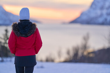 A girl stands on a snowy landscape in Norway, looking at the sunset over the fjord. The scene shows mountains and water under a colorful sky. © Eclipsnet