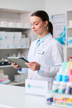 Vertical shot of young pharmacist checking tablet in pharmacy, managing inventory and patient service. Useful for healthcare retail, medication management, digital workflow