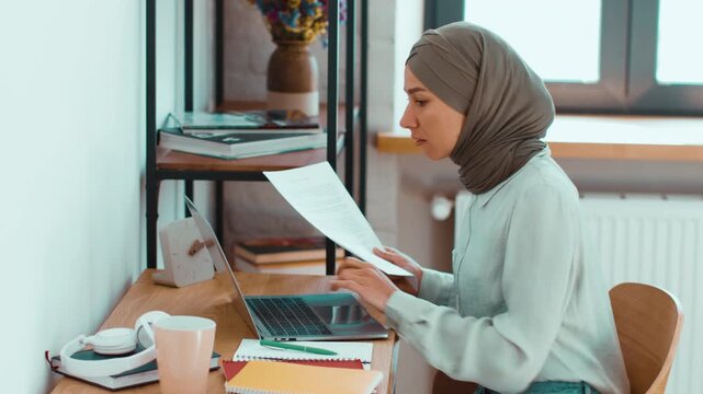A woman with a light skin tone and headscarf is focused on her laptop while holding notes. She sits at a wooden desk with colorful notebooks and a drink nearby.