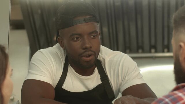 Close view of African American male speaking with customers at window. Man leaning forward and taking order. Wearing apron and cap. Working inside food truck kitchen. Serving couple during shift.