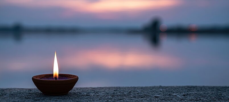 A single lit clay lamp glows on a textured surface against a softly blurred sunset over a tranquil body of water