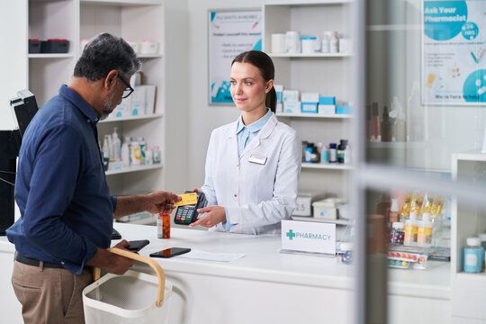 Middle aged man paying by card at pharmacy counter while young female pharmacist processing purchase, illustrating retail healthcare service for pharmacy marketing