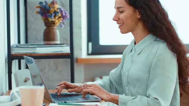A young woman with long curly hair sits at a desk in a modern home office. She uses her laptop while smiling. A vase with flowers sits nearby, creating a productive environment.