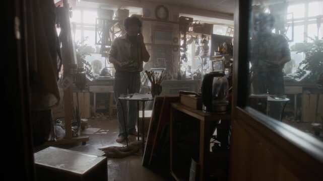Anxious male artist throwing sketches off table, calling on retro landline phone and drinking whiskey in messy workshop during period of creative struggle