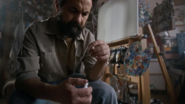 Tilt up shot of miserable male painter sitting in in art studio, holding cigarette and lighter, looking at blank canvas and waiting for inspiration during creative block
