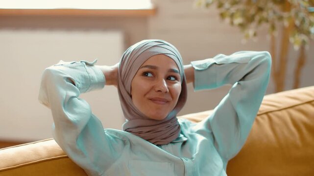 A woman with light skin and a hijab sits comfortably on a yellow sofa. She leans back with her hands behind her head, enjoying a moment of relaxation indoors on a sunny day.